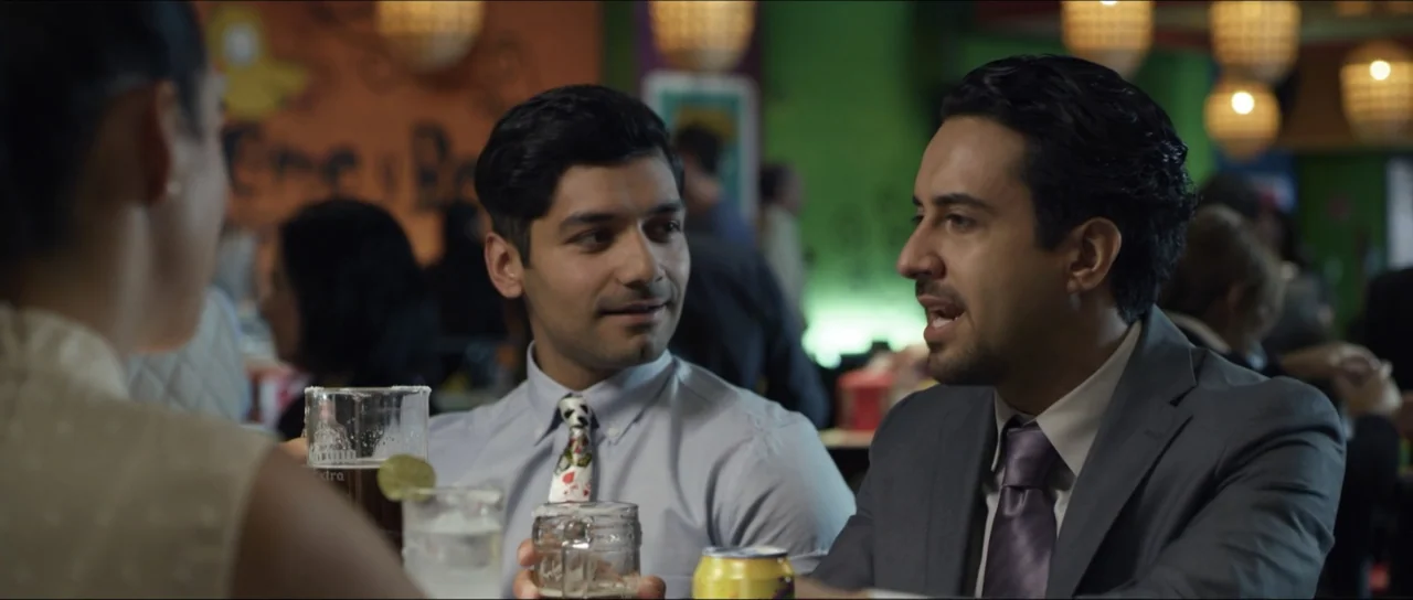 Mirreyes vs Godínez (2019) movie still: medium — A restaurant interior features green walls, hanging basket lights, and blurred diners. Two men in…