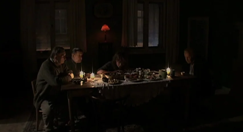 Inland Empire (2006) movie still: wide — A dimly lit room contains four people seated around a large wooden table. They are gathered for a meal at…