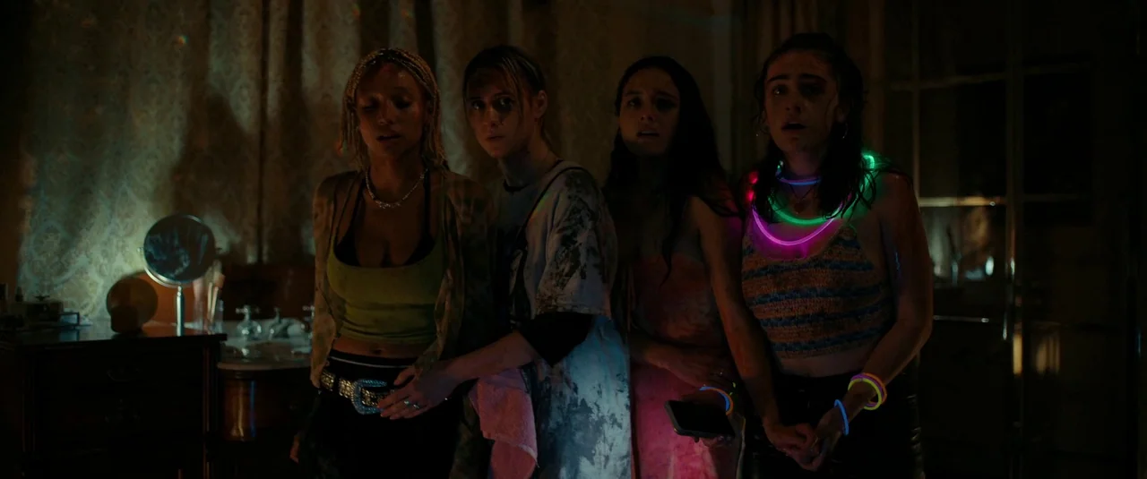 Bodies Bodies Bodies (2022) movie still: medium — A dimly lit room with wallpaper featuring a decorative pattern behind four young women. The four…