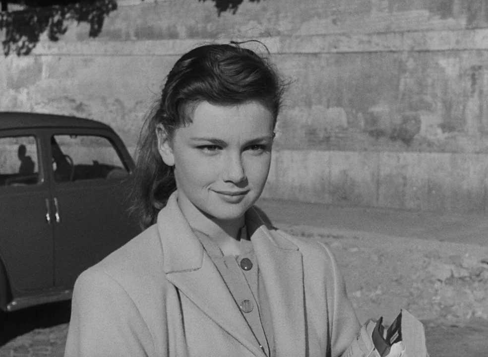 Il Bidone (1955) movie still: medium — An outdoor city sidewalk next to a vintage sedan and a stone wall. A young woman stands looking toward the…