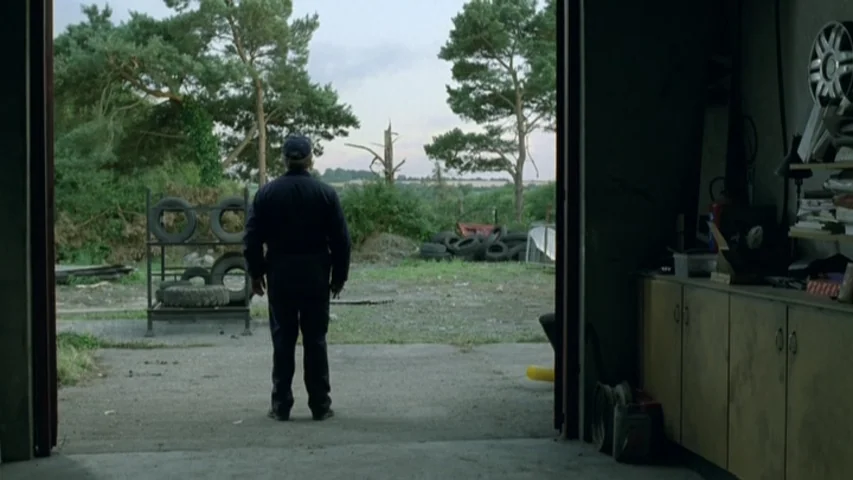 Garage (2007) movie still: fullbody — A cluttered garage interior opens up to a wide outdoor area with trees. A man in a blue jumpsuit stands at the…