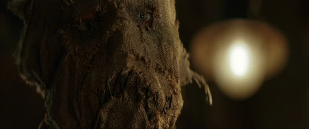 Batman Begins (2005) movie still: closeup — A figure wears a coarse, crudely stitched burlap sack mask in a dark space. A soft out-of-focus light…