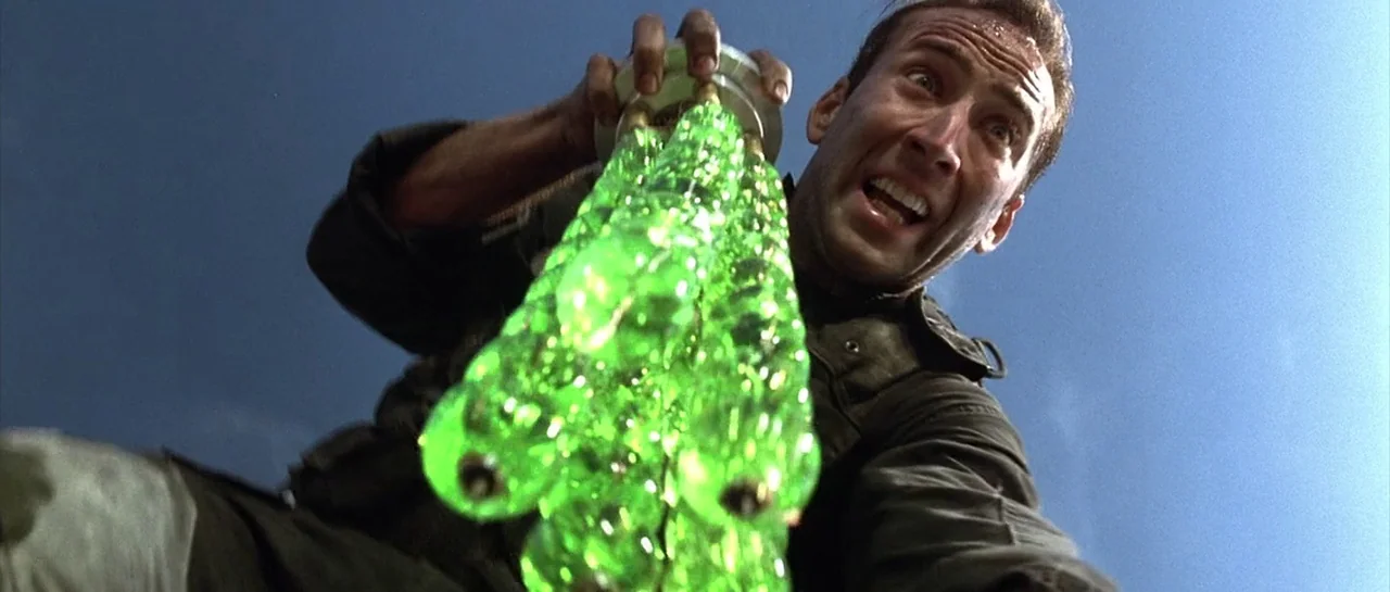 The Rock (1996) movie still: closeup — A man looks down at the camera while holding a glowing green glass sphere cluster. The background is a clear…