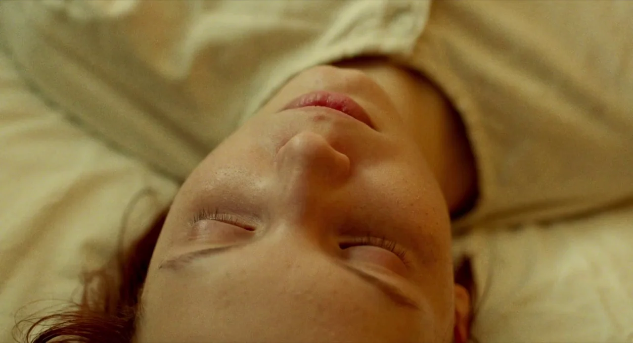 Beanpole (2019) movie still: closeup — A close overhead shot of a young woman resting on white linens. Her head is tilted slightly with her eyes…