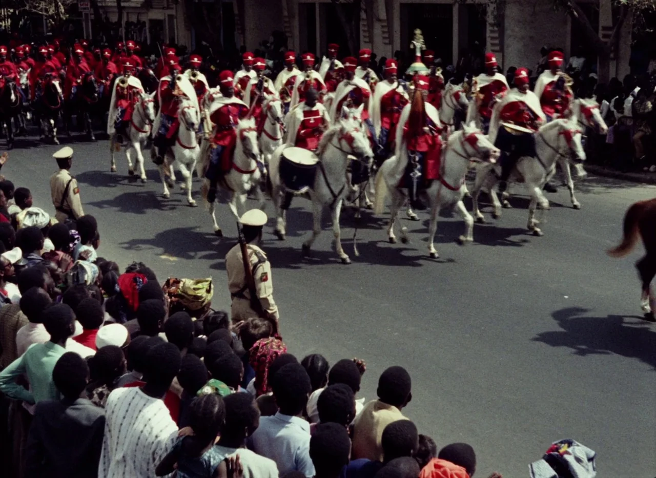 Touki Bouki (1973) movie still: wide — A paved city street is filled with a mounted cavalry unit wearing red coats. A large crowd of spectators…