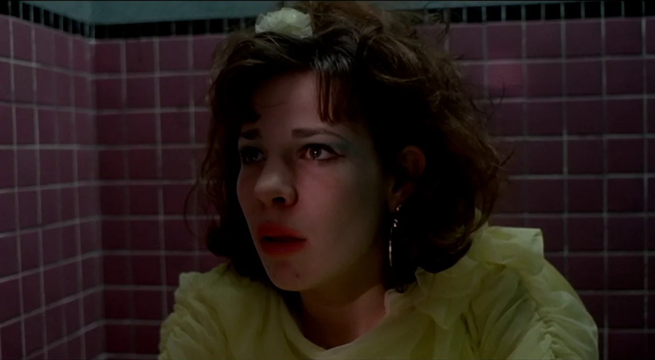 Dogfight (1991) movie still: closeup — A dark, cramped restroom space lined with dark purple square wall tiles. A young woman with dark wavy hair…