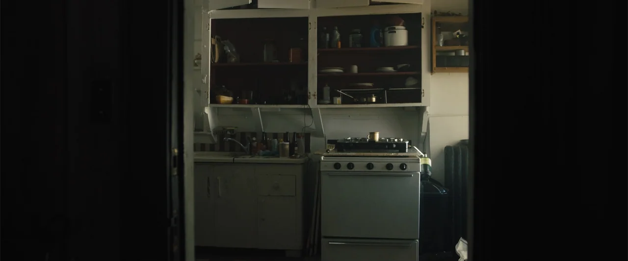 Skin (2009) movie still: medium — A cluttered apartment kitchen with white cabinetry and a vintage gas stove. The room is empty and illuminated by…