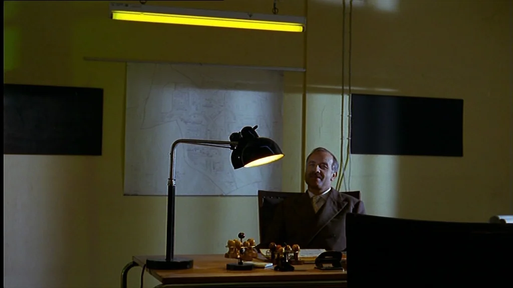 Lola (1981) movie still: medium — A dimly lit room features pale yellow walls with two black rectangular wall panels. A man wearing a suit sits at a…
