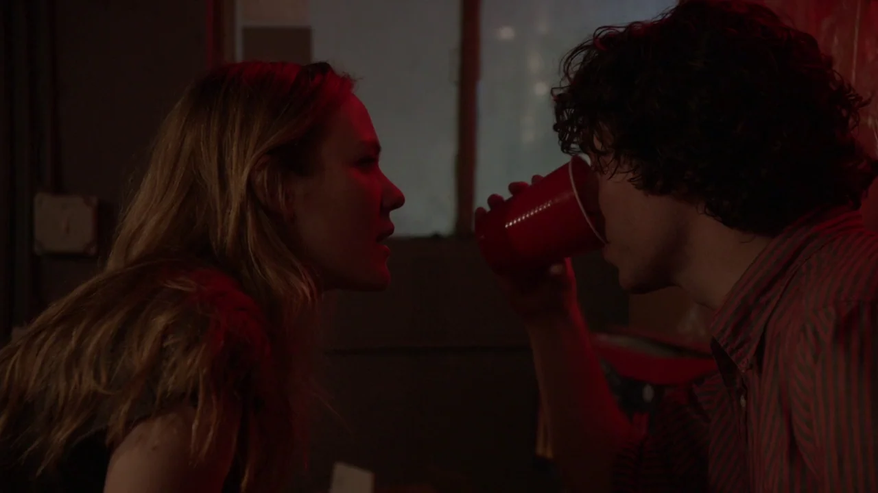 The Heart Machine (2014) movie still: medium — A dimly lit interior room at night features a man and woman in close proximity. The man holds a red…