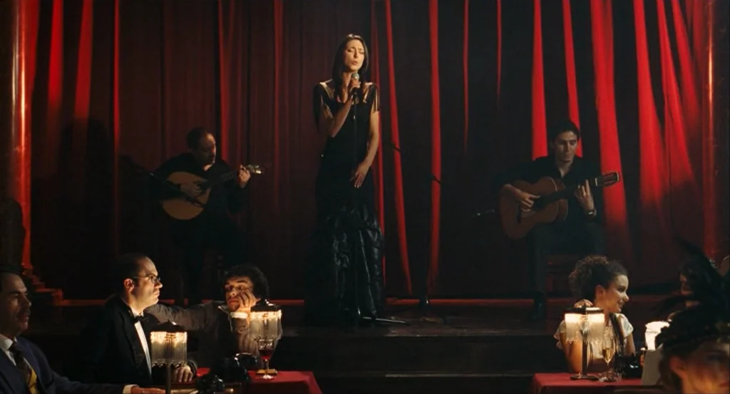 This Night (2008) movie still: wide — A dark supper club features a raised stage with a vibrant red curtain background. A female singer performs on…