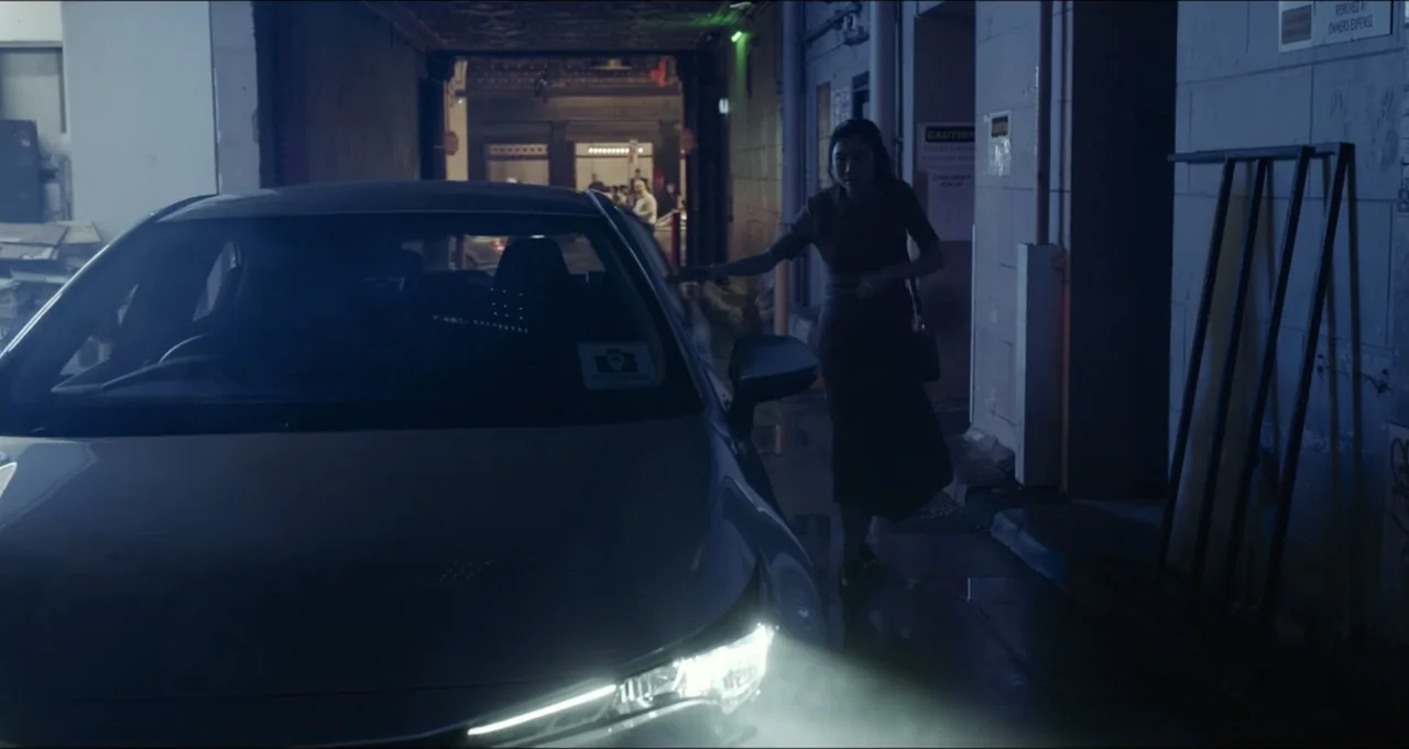 Five Blind Dates (2024) movie still: medium — A narrow urban alleyway at night features dark brick walls and illuminated signs. A woman in dark…