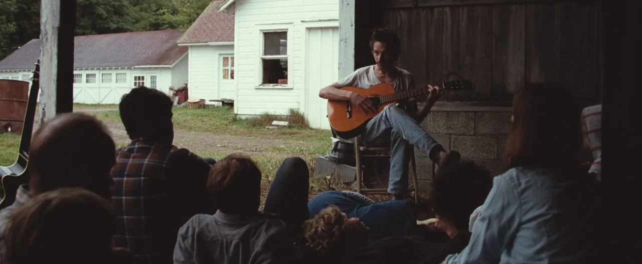 Martha Marcy May Marlene (2011) movie still: wide — A group of people sits inside a dark barn watching a musician perform. The musician sits on a…