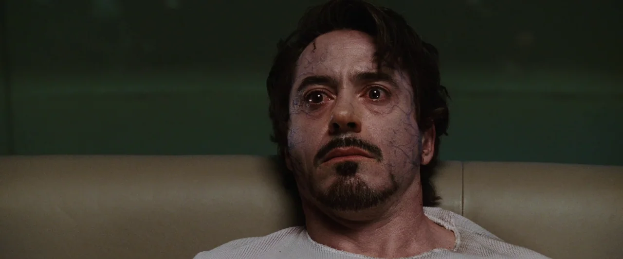 Iron Man (2008) movie still: closeup — A dimly lit room with a muted green wall and a cream-colored headrest. A man with visible blue veins on his…