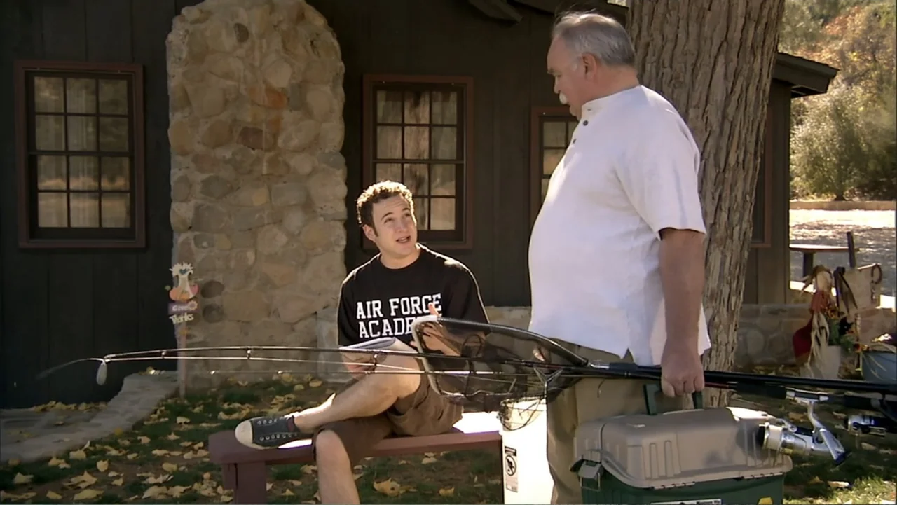 Hot Gimmick: Girl Meets Boy (2019) movie still: medium — A rustic cabin exterior features a stone chimney and wood-paneled walls. A young man sits…
