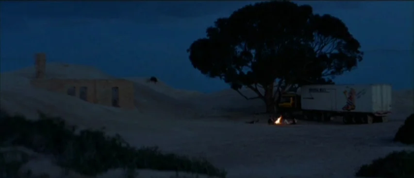 Roadgames (1981) movie still: wide — A vast, dark desert landscape at night features a large tree and a semi-truck. A small campfire glows brightly…