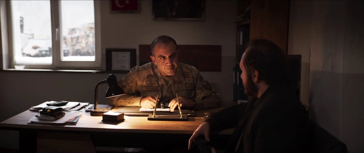 About Dry Grasses (2023) movie still: medium — A dimly lit room features a wooden desk with papers and a lamp. A military officer engages in a…