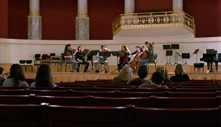 The Piano Teacher (2001) movie still: wide — A spacious concert hall features deep red walls and marble columns behind a stage. A group of musicians…