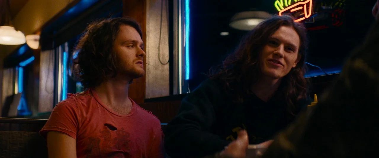 Dinner in America (2020) movie still: medium — Two young men with long brown hair sit inside a diner booth at night. A bright red neon sign hangs on…