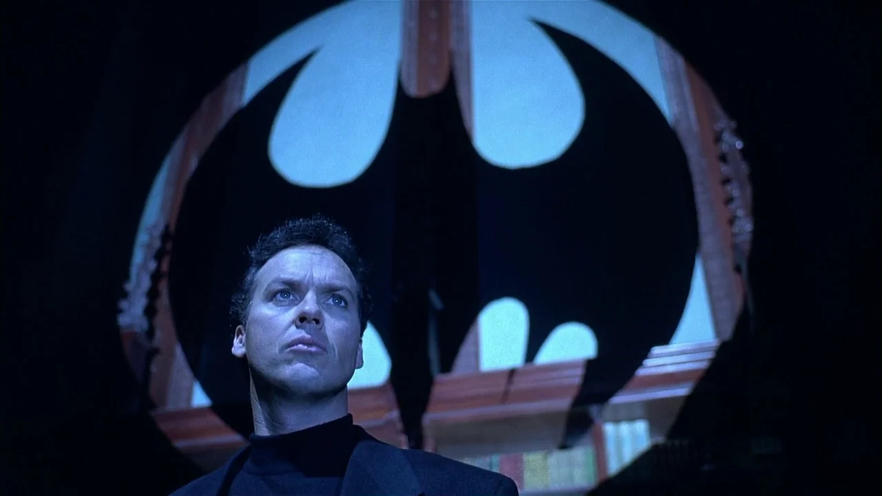 Batman Returns (1992) movie still: medium — A dimly lit room features a large bat-shaped emblem projected onto the back wall. A man in dark clothing…