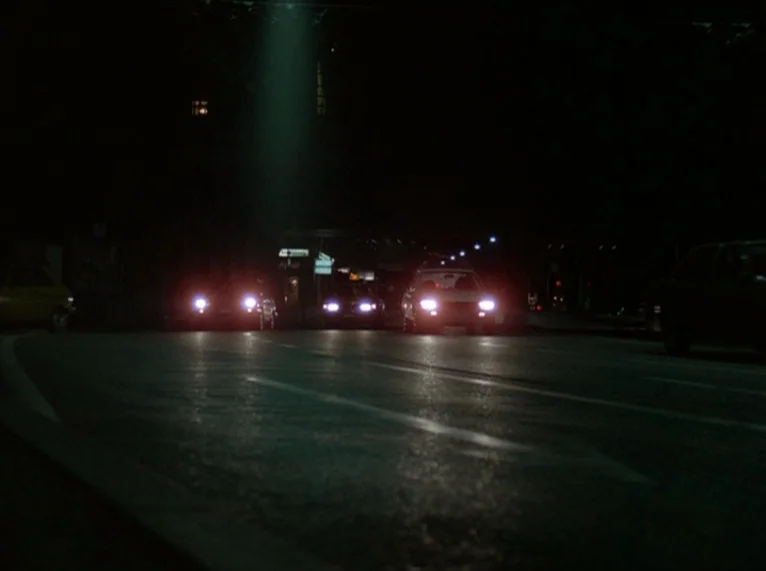 Hail Mary (1985) movie still: wide — An asphalt road at night stretches into the distance under heavy darkness. Several cars drive toward the camera…