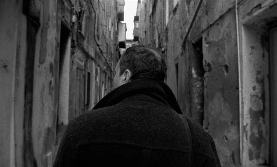 The Man From London (2007) movie still: medium — A narrow stone passageway between two old buildings with weathered, peeling wall surfaces. A man in…