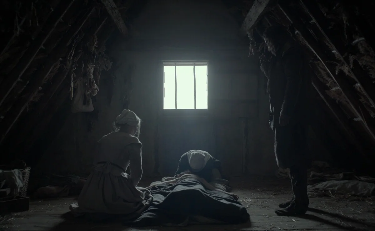 The Witch (2015) movie still: wide — A dimly lit attic with a wooden beamed ceiling and a central window backlight. A man stands while two women…