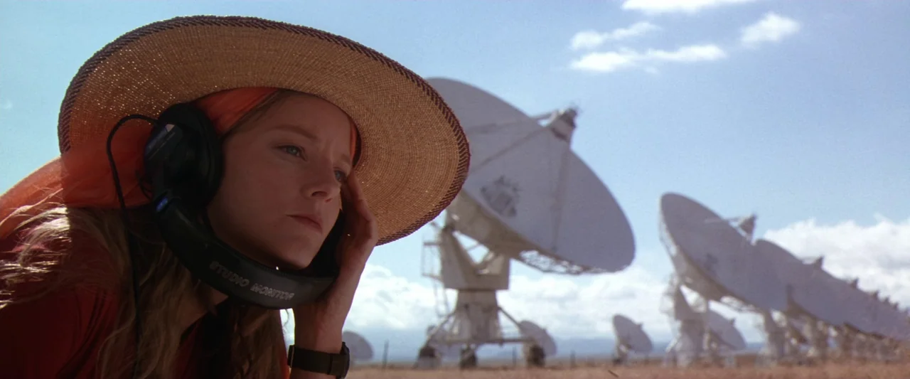 Contact (1997) movie still: closeup — A field filled with large white radio telescope dishes under a blue sky. A woman wearing a straw hat and…