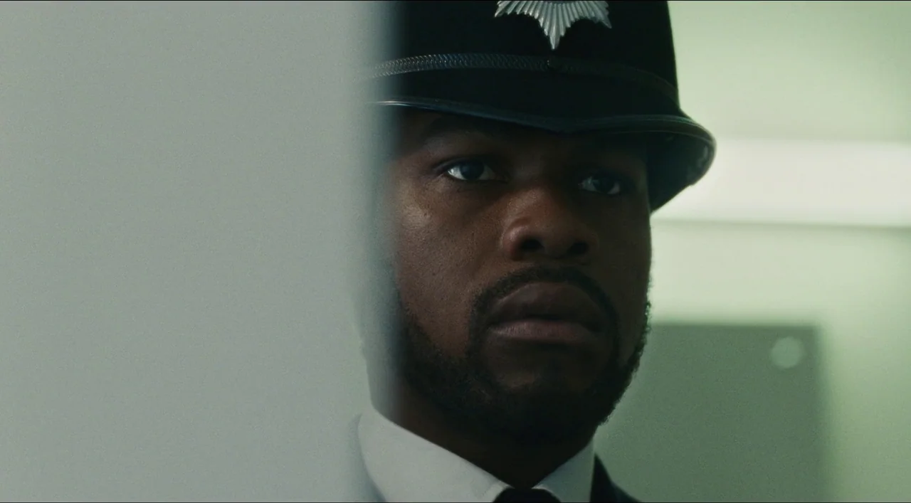 Red, White & Blue (2020) movie still: closeup — A man wearing a British police uniform stands inside a sterile, light-walled room. He looks forward…