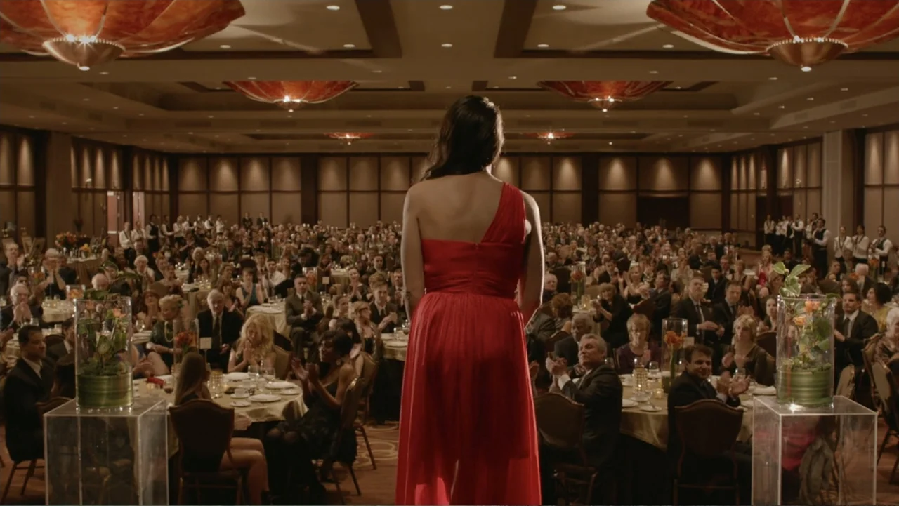 The Captive (2014) movie still: fullbody — A large hotel ballroom is filled with guests seated at circular dining tables. A woman in a red dress…