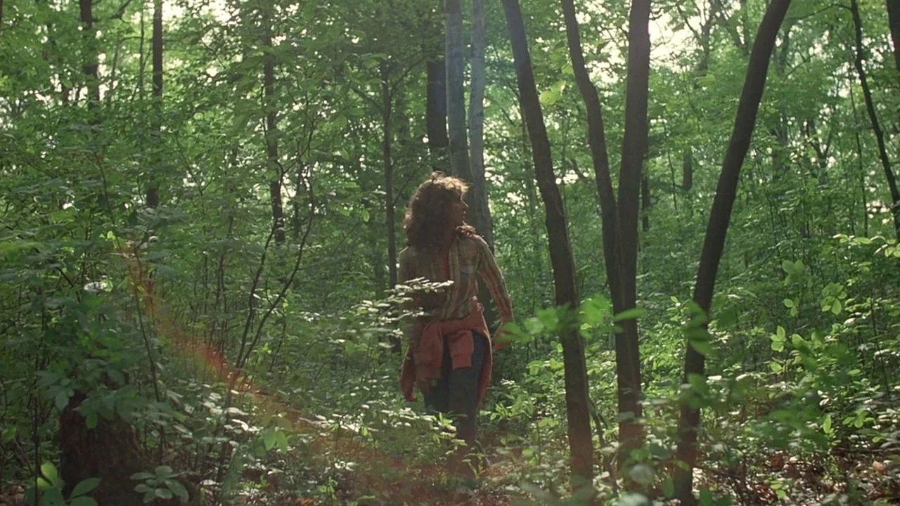 Friday the 13th (1980) movie still: fullbody — A thick forest filled with tall deciduous trees and dense undergrowth of green leaves. A young woman…
