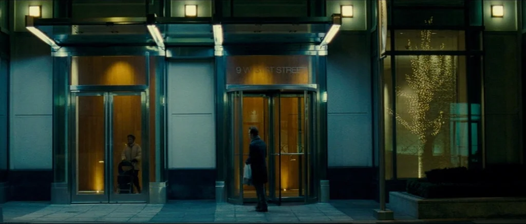 Shame (2011) movie still: wide — A modern building exterior at night with a central revolving door and glass canopy. A man walks into the building…