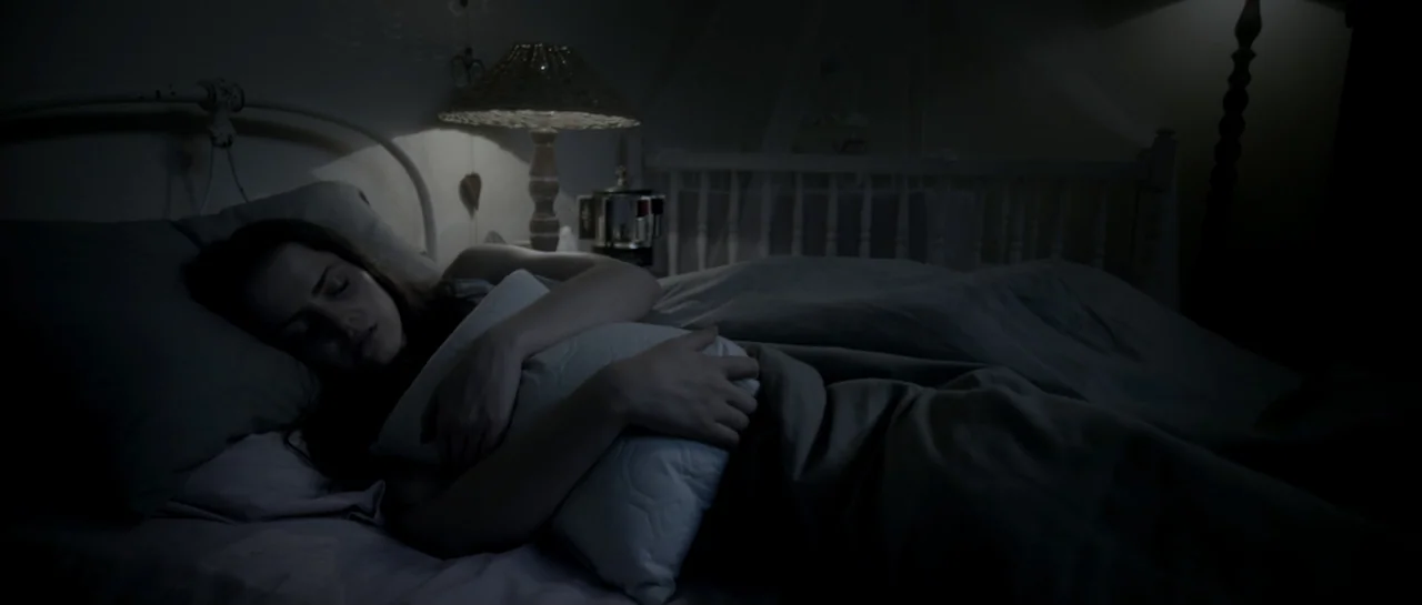 The Lullaby (2019) movie still: medium — A dimly lit bedroom features a white metal bed frame and a lamp. A woman lies under covers while holding a…