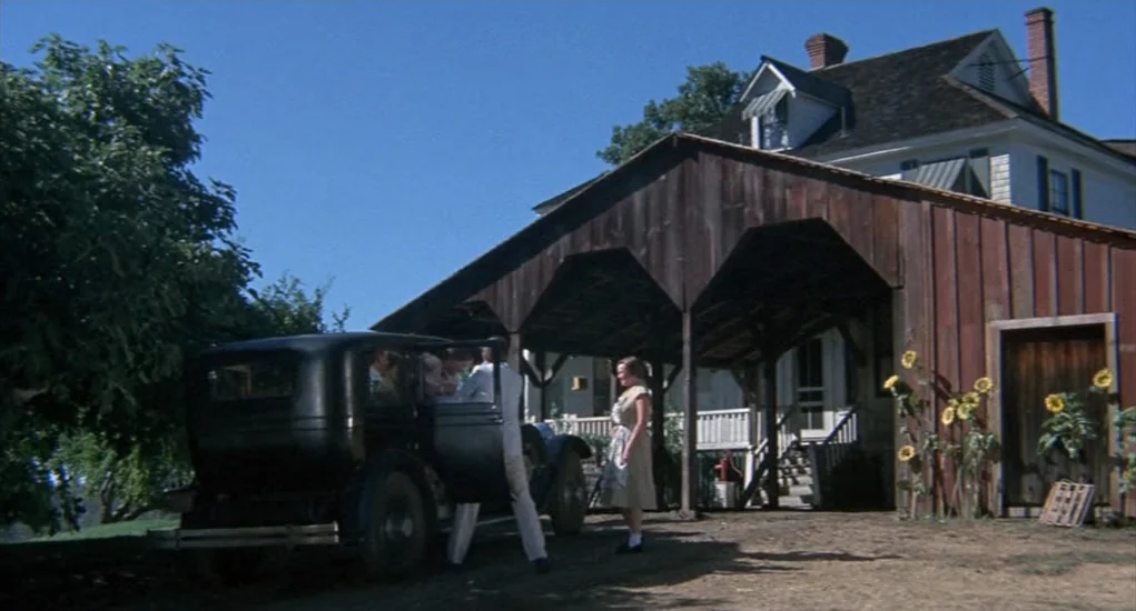 The Other (1972) movie still: wide — A rustic farm yard features a large wooden barn structure next to a house. A man steps out of a dark vintage…
