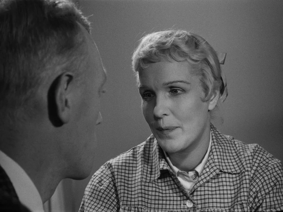 Brink of Life (1958) movie still: medium — Two individuals sit in a nondescript indoor space with a plain, neutral background wall. A woman in a…