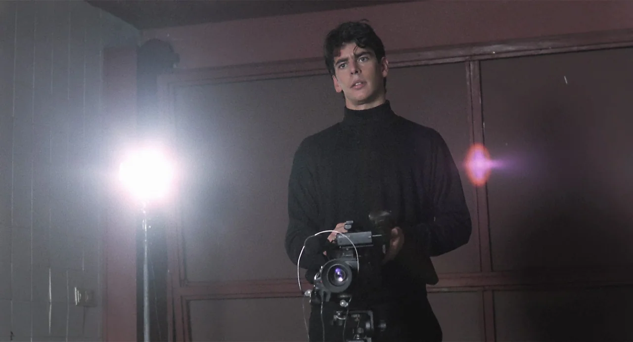 Thesis (1996) movie still: medium — An interior space features wood-paneled walls and a bright light source on a stand. A young man wearing a black…
