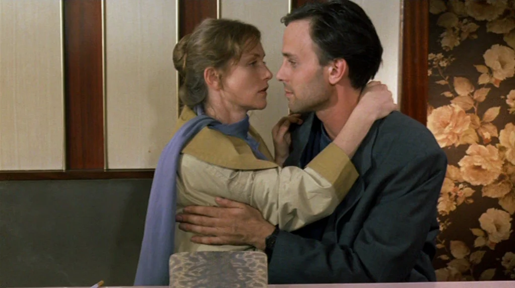 Malina (1991) movie still: medium — The interior features walls covered with beige vertical panels and vintage floral wallpaper. A woman and a man…