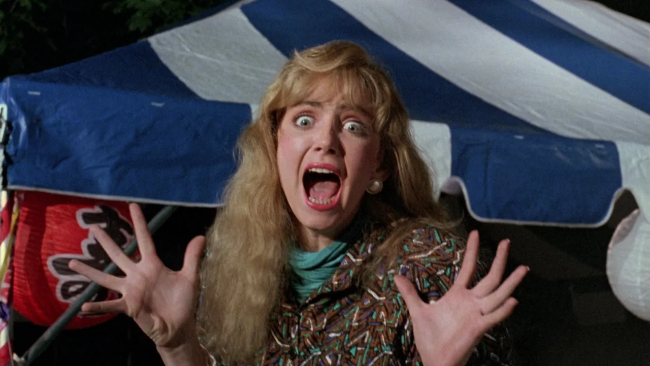 Frankenhooker (1990) movie still: medium — A dark outdoor carnival scene with blue and white striped tent canopy visible behind. A young blonde…