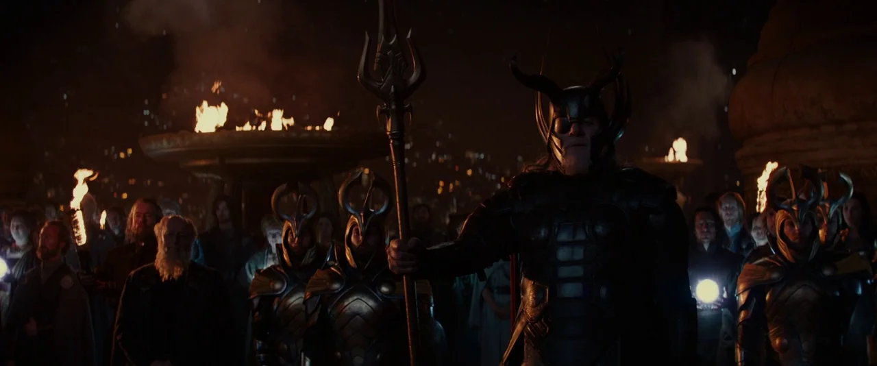 Thor: The Dark World (2013) movie still: wide — A dark, torch-lit ceremonial plaza at night features a crowd gathered in the gloom. An armored king…
