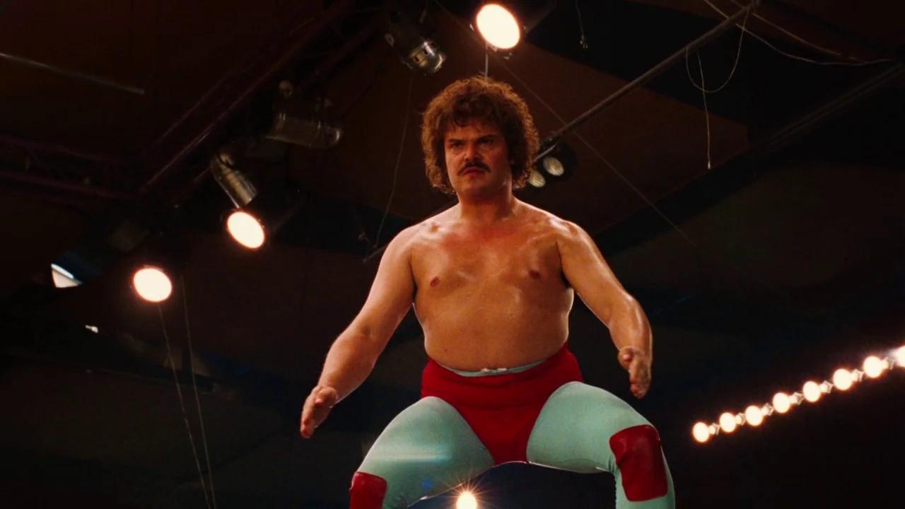 Nacho Libre (2006) movie still: medium — A man in wrestling attire stands in the center of a dark indoor arena. He is poised in a crouched stance…