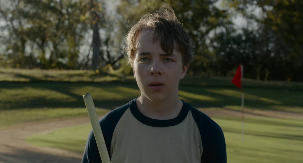 Wildlife (2018) movie still: medium — A teenage boy stands on a putting green with trees in the background. He holds a flagstick while looking…