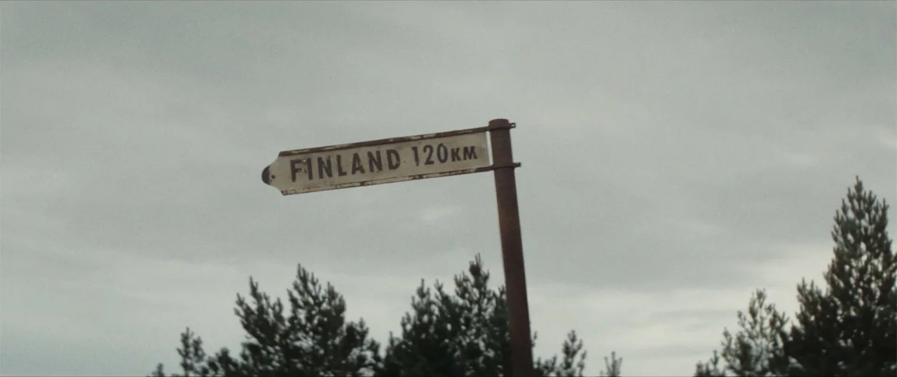 Sisu: Road to Revenge (2025) movie still: medium — An aged metal road sign stands on a post against a thick, overcast sky. Tall pine trees are…