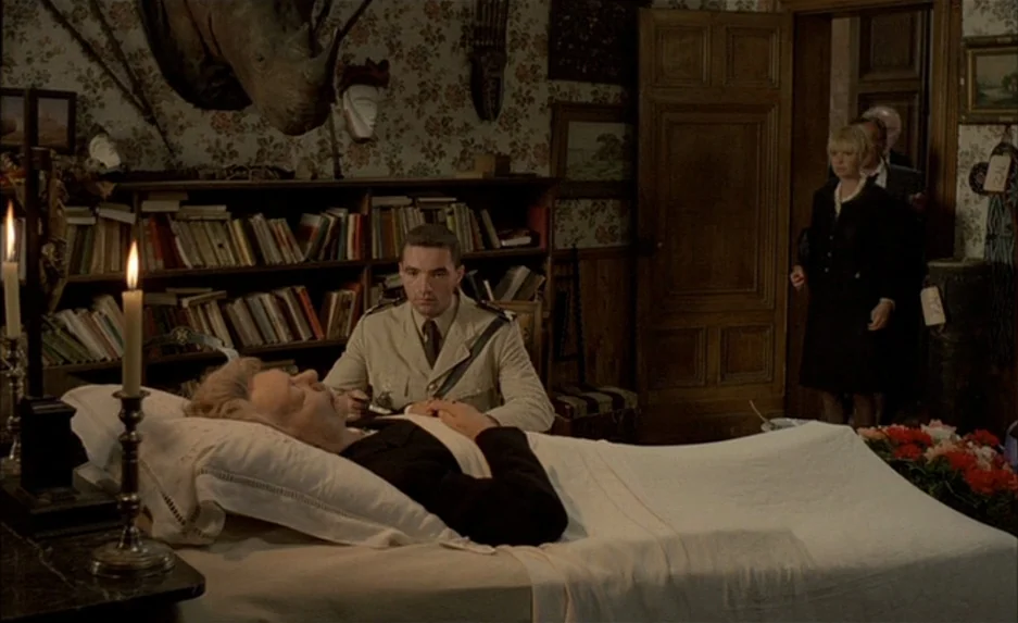 Milou en Mai (1990) movie still: medium — A room with patterned floral wallpaper contains a bookshelf and an animal head mount. A person rests in…