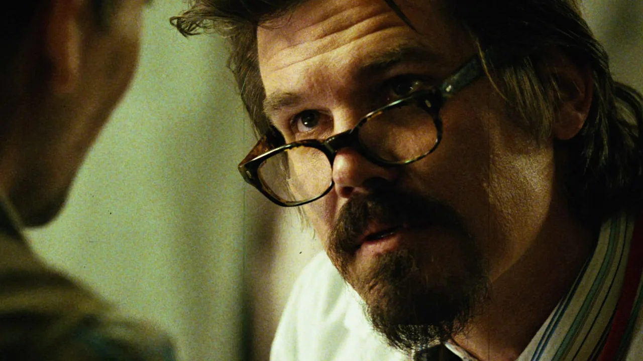 Planet Terror (2007) movie still: closeup — An interior space features muted walls and clinical, dim lighting. A man wearing thick-rimmed glasses…