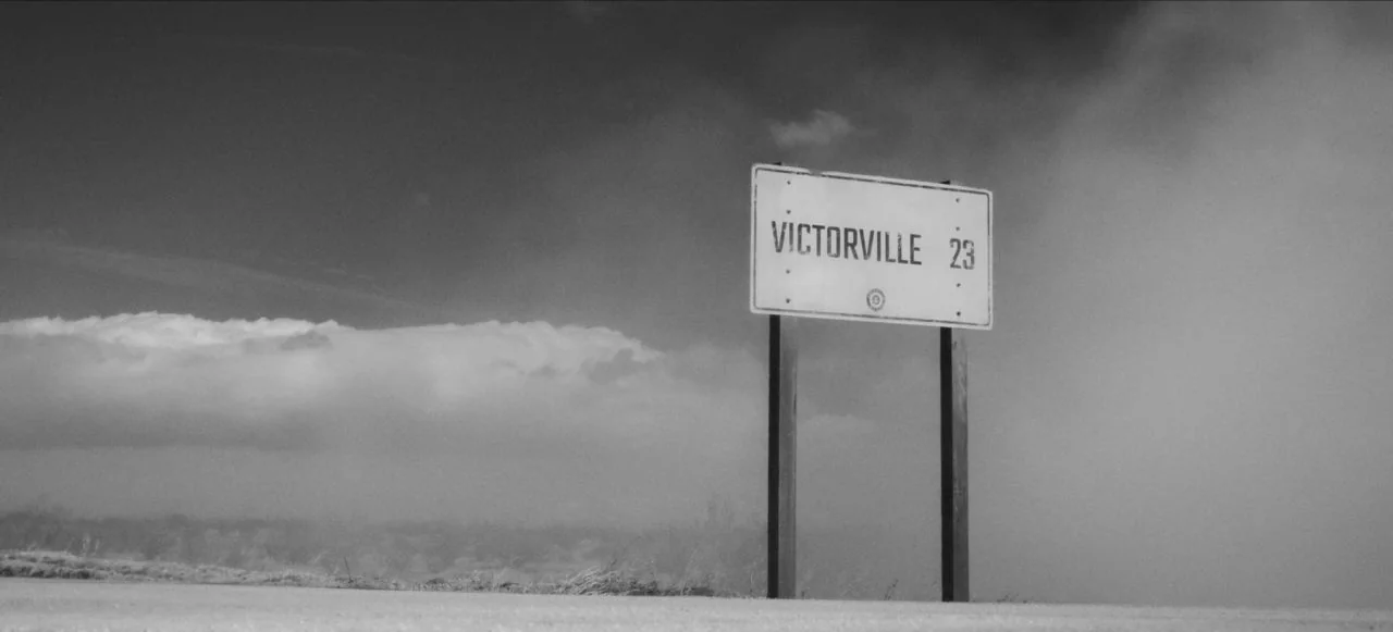 Mank (2020) movie still: wide — A metal road sign stands against a desolate landscape under a heavy cloudy sky. The sign displays text indicating a…