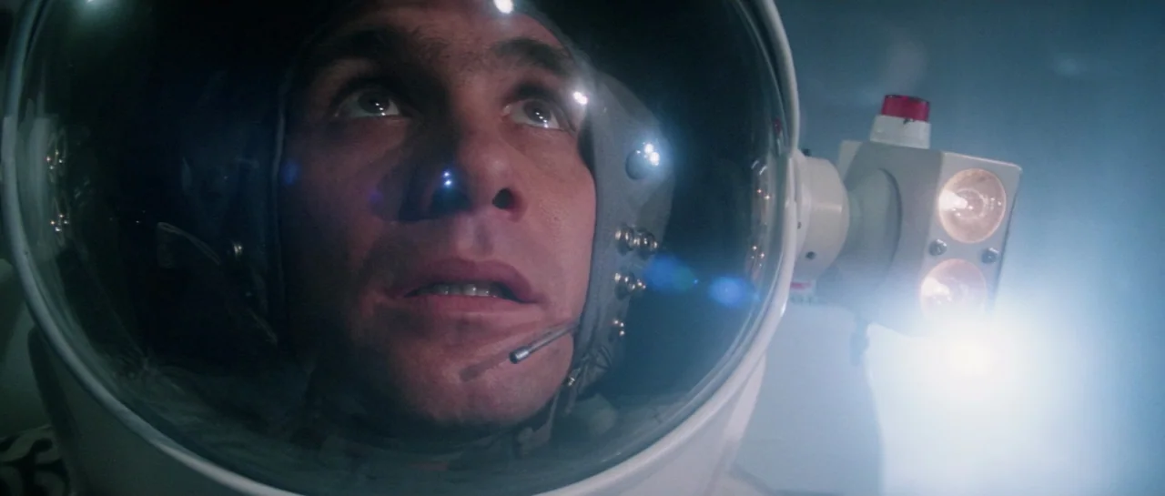 Lifeforce (1985) movie still: closeup — An astronaut in a white space helmet is lit by a mounted spotlight. The subject looks upward into a dark…