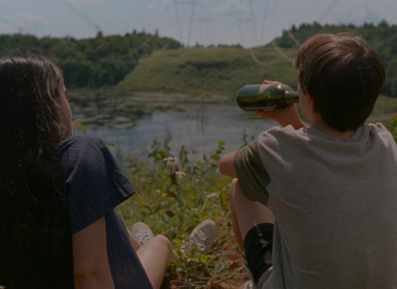 Falcon Lake (2022) movie still: medium — A grassy hill overlooks a murky pond surrounded by green vegetation and trees. A young male and female sit…