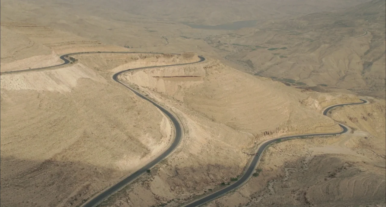 Incendies movie still: establishing — A winding asphalt road cuts through a vast, barren desert mountain range. The terrain features rolling sandy…