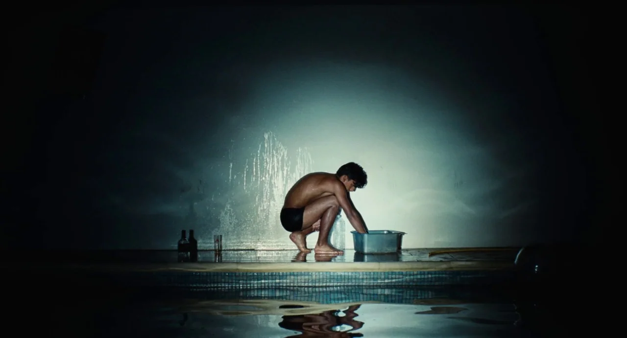 Ava (2017) movie still: wide — A dark poolside edge with a damp concrete wall covered in water streaks. A shirtless man crouches beside a metal…