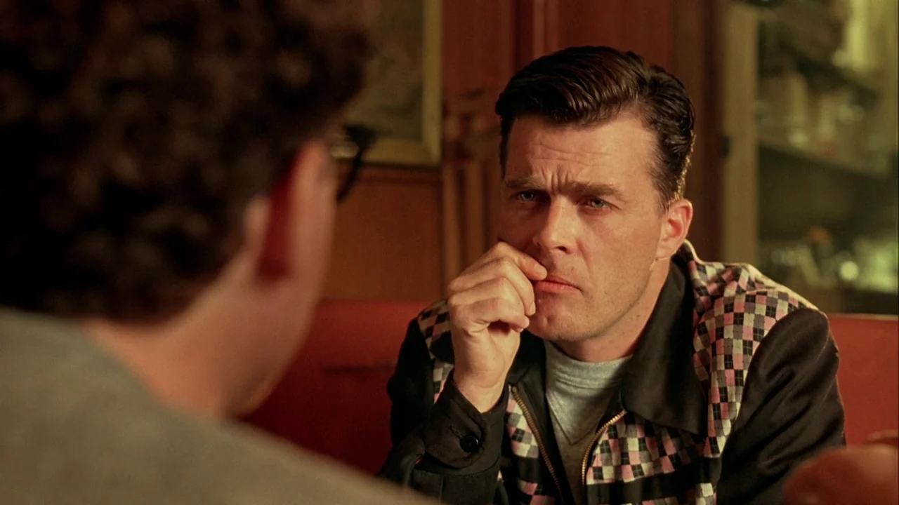 Naked Lunch (1991) movie still: medium — A dimly lit interior with reddish walls and a vintage diner aesthetic. A man with dark styled hair sits…