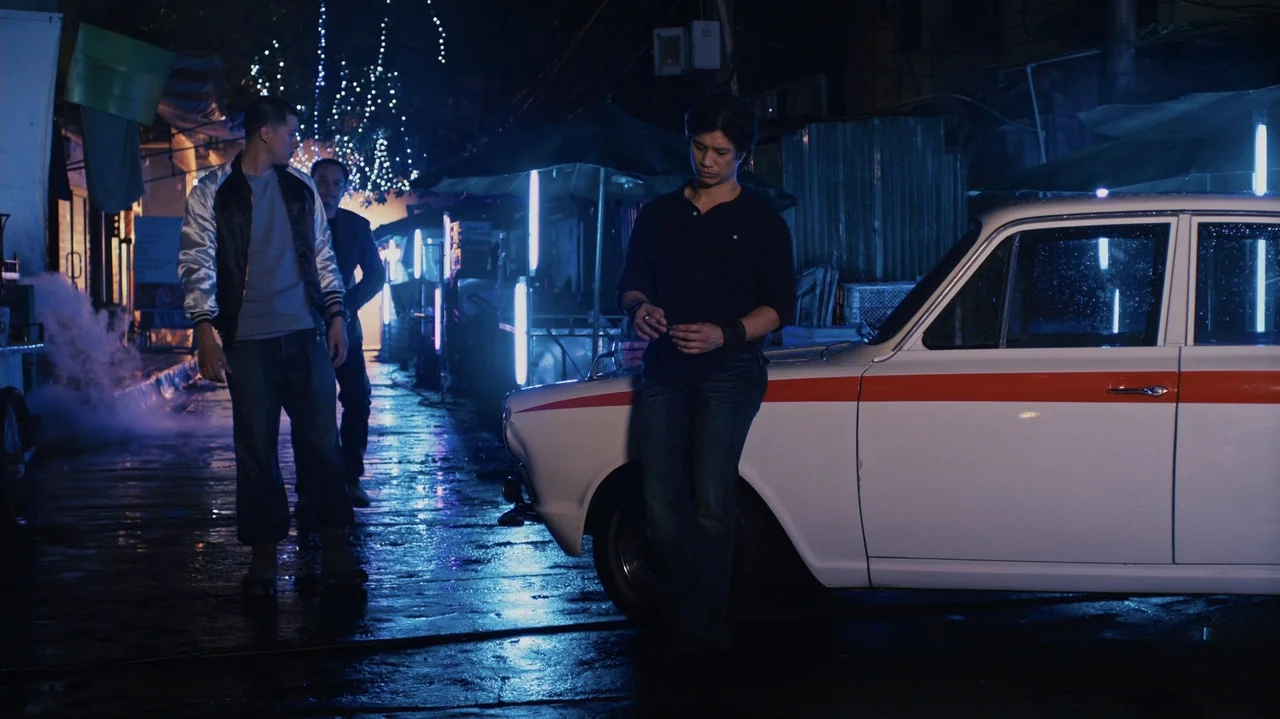 Zero Tolerance (2015) movie still: medium — A wet asphalt street at night features a white vintage sedan with a red stripe. A man stands beside the…